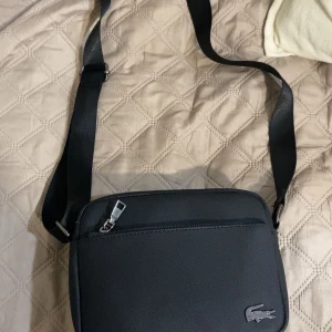 Svart axelväska från Lacoste - Snygg svart axelväska från Lacoste med klassisk krokodil-logga i metall på framsidan. Väskan har justerbar rem, flera dragkedjefack och är tillverkad i slitstark syntet. Perfekt storlek för mobil, plånbok och andra essentials.