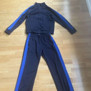 Svart och blå träningsdress - Snygg svart träningsdress med blå ränder längs ärmar och ben. Jackan har dragkedja och ståkrage, byxorna har resår i midjan för skön passform. Perfekt för dig som gillar sportig stil.