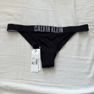 Svart bikiniunderdel Calvin Klein M - Svart bikiniunderdel från Calvin Klein med bred resår i midjan och tydlig logga framtill. Helt oanvända då dem ej passade mig.   Snygg, enkel design i stretchigt material som sitter skönt på kroppen. Perfekt för strand eller poolhäng. Materialet är syntet och modellen är klassisk med hög benskärning.