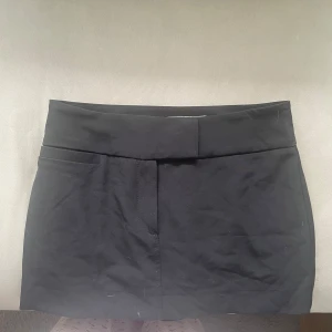 Svart kortkjol med bred midja - Enkel och stilren svart kortkjol med bred midjeresår och inbyggda shorts. Kjolen har en rak passform och är perfekt för dig som gillar minimalistisk stil. Passar till många olika outfits och är lätt att matcha med både sneakers och boots.