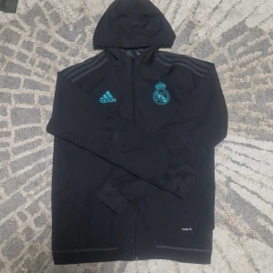 Real Madrid vindjacka - Svart vindjacka från Adidas med Real Madrid-logga och blåa detaljer. Jackan har huva, hel dragkedja och klassiska Adidas-ränder på axlarna. Tillverkad i lätt polyester med Climalite-teknologi, perfekt för fotbollsträning eller chill vardagsstil.
