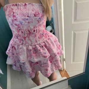 Rosa blommig volangklänning - Trendig klänning🤩Skulle säga att den passar som S/M!💗 Supersöt kort klänning i rosa och lila toner med blommigt mönster. Axelbandslös modell med smock upptill och flera volanger i kjolen. Tunt, lätt material som känns perfekt för varma dagar. Söt spetskant längs volangerna för extra detalj.