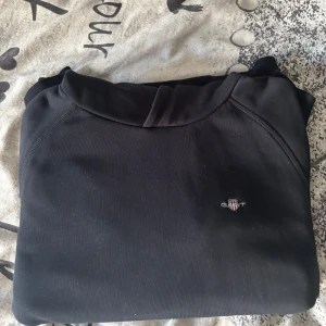 Svart hoodie från GANT med logga - Svart hoodie från GANT med broderad logga på bröstet. Klassisk känguruficka framtill och huva. Tillverkad i mjuk bomullsmix, perfekt för chill dagar. Ribbade muddar vid ärmslut och nederkant för en snygg passform. Inga hål och inga fläckar. FRI FRAKT