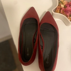 Vinröda ballerinaskor från Lindex - Snygga ballerinaskor från Lindex i vinröd mocka-look. Skorna har spetsig tå och platt sula, vilket ger en trendig och stilren vibe. Perfekta för dig som vill ha en elegant touch till outfiten. Färg och material gör dem lätta att matcha.