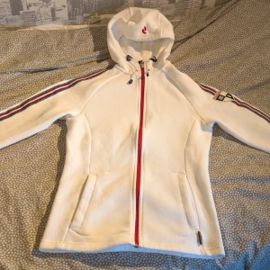 Vit fleecejacka Peak Performance M - Vit fleecejacka från Peak Performance med dragkedja i rött. Snygga ränder i rött, blått och vitt längs ärmarna samt broderad logga på ena ärmen och vid huvan. Jackan har två fickor framtill och är tillverkad i mjuk polyester.