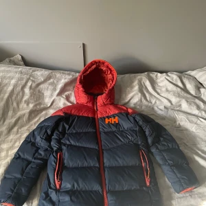 Röd och marinblå pufferjacka Helly Hansen - Snygg pufferjacka från Helly Hansen i rött och marinblått med orange detaljer. Jackan har huva, dragkedja framtill och två fickor med dragkedja. Fodret är rött och loggan HH syns tydligt på bröstet och Helly Hansen på ryggen. Perfekt för kalla dagar.