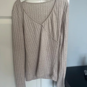Beige ribbad omlottopp - Säljer en beige ribbad topp med omlottdetalj och v-ringning. Långärmad och figurnära passform, perfekt till jeans eller kjol. Mjuk och stretchig, superenkel att styla för en chill look.