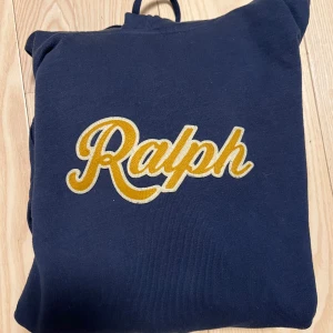 Polo Ralph Lauren hoodie - Snygg mörkblå hoodie från Polo Ralph Lauren med broderad gul text 'Ralph' på bröstet. Klassisk känguruficka framtill, dragsko i huvan och mjukt bomullsmaterial. Perfekt för en chill och stilren look. Nypris 2700kr