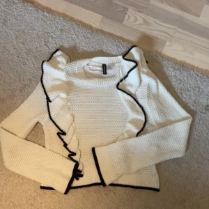 Vit stickad volangtröja H&M Divided XS - Superfin vit stickad tröja från H&M Divided i storlek XS. Tröjan har långa ärmar, volangdetaljer framtill och svarta kontrastsömmar längs volanger och kanter. Perfekt för dig som gillar en söt och trendig look.