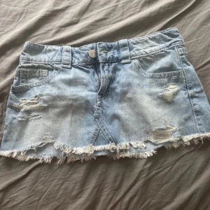 Ljusblå jeanskjol från Gina Tricot - Säljer en ljusblå jeanskjol från Gina Tricot med slitna detaljer och fransig nederkant. Kjolen har klassiska fickor fram och bak samt bälteshällor. Perfekt för dig som gillar en avslappnad och trendig stil.
