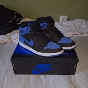 Nike Air Jordan 1 High Royal Reimagined - Säljer ett par Nike Air Jordan 1 High OG i svart och blå mocka med klassisk vit sula. Skorna har snörning, rund tå och ikoniska Jordan Wings-loggan på sidan. Perfekta för dig som vill sticka ut med en tidlös sneaker i snygga färger. Box medföljer👌🏻