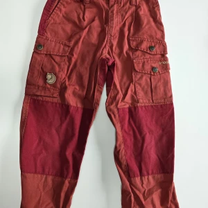 Röda cargobyxor från Fjällräven barn - Snygga röda cargobyxor från Fjällräven med förstärkta knän och flera praktiska fickor med knappar. Byxorna har klassisk logga på benet och är tillverkade i slitstark bomull. Perfekta för äventyr utomhus och har cool tvåtonad design.