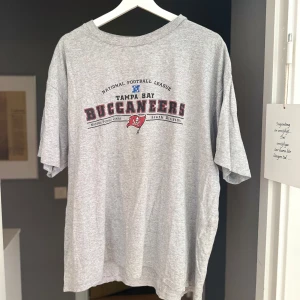 Grå Tampa Bay Buccaneers t-shirt - Snygg grå t-shirt med Tampa Bay Buccaneers-tryck framtill. Oversized passform och klassisk crewneck. Perfekt för dig som gillar amerikansk fotboll och vill ha en chill och sportig stil. Sitter som L