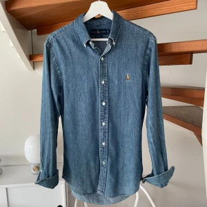 Ralph Lauren Jeansskjorta - Snygg jeansskjorta från Ralph Lauren i tvättad blå denim. Slim fit-modell med klassisk krage, knäppning framtill och broderad logga på bröstet. Långärmad och perfekt för en avslappnad men stilren look.