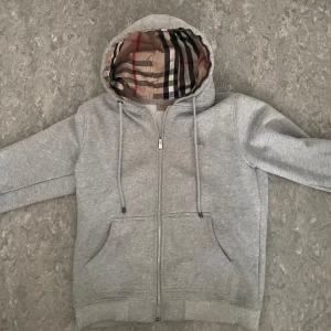 Grå hoodie från Burberry med dragkedja - Snygg grå hoodie från Burberry med klassiskt rutigt foder i huvan och broderad logga på bröstet. Tröjan har dragkedja, snörning i huvan och två fickor framtill. Perfekt för dig som gillar stilrena och exklusiva plagg.