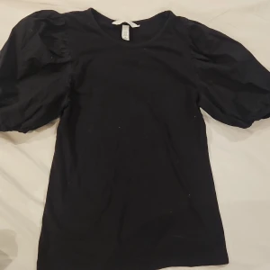Svart topp med puffärmar från H&M - Svart topp från H&M i storlek XS med trendiga puffärmar som ger en cool siluett. Tillverkad i mjuk bomull med lite elastan för extra komfort. Perfekt för dig som gillar en enkel men ändå statement look.