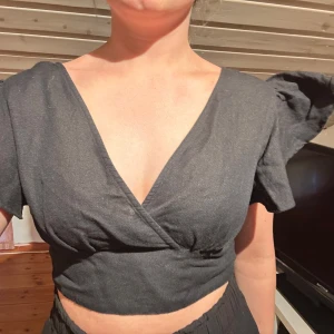 Svart omlottopp från BikBok - Svart croppad omlottopp från BikBok med v-ringning och korta volangärmar. Toppen har öppen rygg med en smal rem upptill och sitter tajt i midjan. Perfekt för dig som gillar trendiga och stilrena plagg med snygga detaljer.