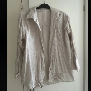 Randig beige skjorta med krage - Snygg beige skjorta med tunna vita ränder och klassisk krage. Skjortan har lång ärm och bröstficka, perfekt att bära öppen eller stängd. 