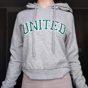 Grå hoodie UNITED från FB Sister - Grå hoodie från FB Sister i storlek S med grönt och vitt UNITED-tryck på bröstet. Klassisk huva med snörning och långa ärmar. Mjuk och skön bomullsblandning, perfekt för chill dagar eller när du vill ha en avslappnad stil.