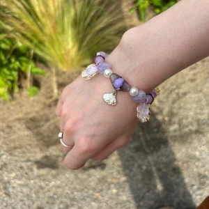 Lila pärlarmband med snäckberlocker - Superfint armband med lila och vita pärlor, mixade former och små blommor. Utsmyckat med guldiga snäckberlocker som ger en unik och somrig vibe. Perfekt accessoar för dig som vill sticka ut med något färgglatt och lekfullt.
