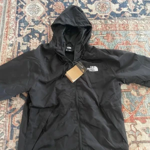  vindjacka från The North Face  - Svart vindjacka från The North Face med huva och dragsko. Jackan har dragkedja framtill, två sidofickor och vit broderad logga på bröstet. Lätt och smidig modell i syntetmaterial, perfekt för blåsiga dagar.