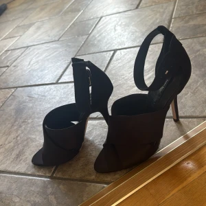 Svarta klackar från Zara Basic - Snygga svarta klackar från Zara Basic med breda korslagda band över foten och öppen tå. Skorna har hög klack och justerbart ankelband med spänne. Tillverkade i ett matt tygmaterial som ger en stilren look.