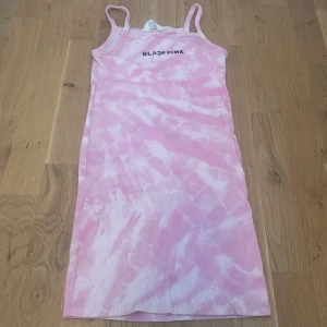 Rosa tie-dye klänning BLACKPINK H&M - Snygg ribbad kortklänning från H&M med rosa och vit tie-dye mönster och trycket BLACKPINK framtill. Klänningen är ärmlös med smala axelband och har en slim passform. Perfekt för dig som älskar K-pop och vill sticka ut med en cool look.