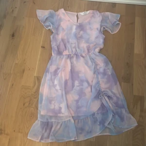 Pastellfärgad omlottklänning H&M - Superfin kortklänning från H&M i pastelliga toner av lila, rosa och blått. Klänningen har omlottkänsla med volangkant nertill, korta vingar till ärmar och knyt i sidan. Lätt och luftig, perfekt för varma dagar.