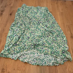 Blommig långkjol från Lindex - Söt långkjol från Lindex med blommigt mönster i grönt, lila, vitt och gult. Kjolen har resår i midjan och är tillverkad i ett lätt och luftigt material, perfekt för varma dagar. Volangdetaljer nertill ger en extra touch.