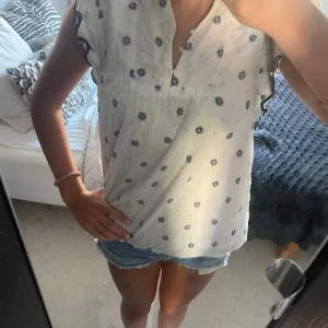 Vit blommig blus från Lindex - Supersöt vit blus från Lindex med små mörkblå blommor och volangdetaljer längs ärmarna. Blusen har v-ringning med knappar och är ärmlös med vågiga kanter. Passar xs-m