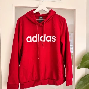 Röd Adidas hoodie med logga - Riktigt snygg röd hoodie från Adidas med stor vit logga framtill. Klassisk huva med snörning och långa ärmar. Mjuk och skön i bomullsmix, enbart använd hemma fåtal gånger