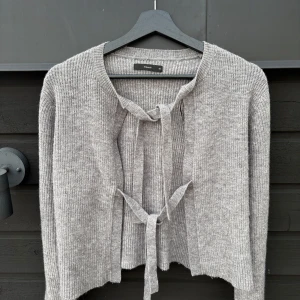 Grå Pigalle Kofta Ny - Superfin grå kofta Stickad modell i mjukt, stretchigt material med två dekorativa knytband framtill. Perfekt att styla över ett linne eller klänning – både vardag och fest! Väldigt bra skick, så gott som ny, endast använd en gång✨ Storlek: XS