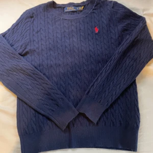 Marinblå kabelstickad tröja Polo Ralph Lauren - Kabelstickad marinblå tröja från Polo Ralph Lauren med klassisk röd broderad logga på bröstet. Tröjan är långärmad, har rund halsringning och är gjord i mjuk bomull. Perfekt för dig som gillar stilren och tidlös design.