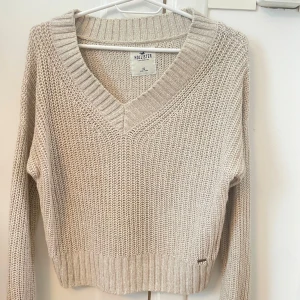 Beige stickad v-ringad Hollister tröja - Supermysig beige stickad tröja från Hollister med v-ringning och ribbade muddar. Långärmad och lite loose fit, perfekt att slänga på sig över ett linne. Passar både till jeans och kjol för en chill look.