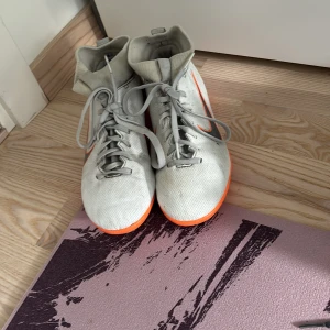 Nike Mercurial vita och orange fotbollsskor - Nike Mercurial fotbollsskor i vitt med grå detaljer och orange sula. Skorna har snörning, högt skaft och är tillverkade i syntetmaterial. Perfekta för dig som vill sticka ut på planen med snygg design och bra grepp.