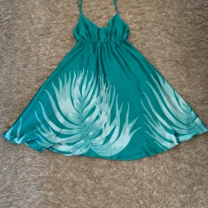 Turkos kortklänning med palmtryck - Snygg turkos kortklänning med smala axelband och v-ringning. Klänningen har ett tropiskt palmbladsmönster i vitt och ljusblått nertill och är gjord i ett glansigt, satinliknande material. Perfekt för sommarens varma dagar och strandhäng.