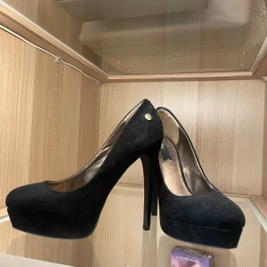Svarta pumps med hög klack från Belle - Snygga svarta pumps från Belle med hög klack och plattform. Skorna har rund tå och är klädda i mockaliknande material som ger en lyxig känsla. Perfekta för dig som vill ha extra höjd och en klassisk look. Något trötta i materialet.