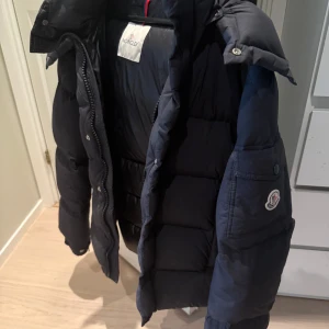 Svart Moncler dunjacka med dragkedja - Säljer en svart dunjacka från Moncler med klassisk puffer-design. Jackan har hög krage, dragkedja och tryckknappar framtill samt Moncler-logga på ärmen. Insidan har ett coolt serietryck och jackan är perfekt för kalla dagar. passar medium perfekt 