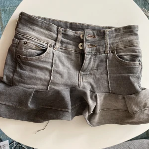 Grå jeansshorts med volang från Bershka - Snygga grå jeansshorts från Bershka med hög midja och dubbla knappar framtill. Shortsen har volangdetalj längst ner, klassiska fickor fram och bak samt cool tvättad look. Perfekta för dig som vill sticka ut lite extra i sommar!