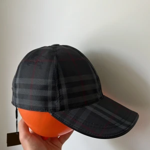 Rutig keps från Burberry i svart - Snygg keps från Burberry i klassiskt rutigt mönster med svarta, gråa och röda toner. Kepsen har böjd skärm, justerbar rem baktill med Burberry-logga och är tillverkad i slitstark polyester. Perfekt accessoar för en trendig streetstyle-look. Pris kan diskuteras.