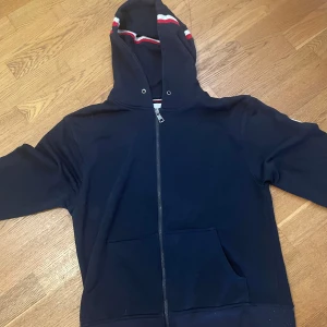 Mörkblå Moncler hoodie med dragkedja - Snygg mörkblå hoodie från Moncler med hel dragkedja, huva och fickor framtill. Ärmarna har vita och röda ränder samt Moncler-logga på ena ärmen. Perfekt för en chill och sportig stil.