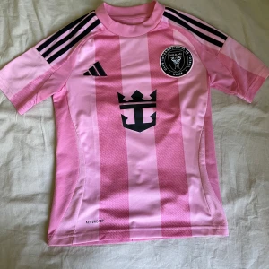 Inter Miami Messi 10 fotbollströja rosa - Säljer en officiell Inter Miami fotbollströja från Adidas i rosa med svarta detaljer. Tröjan har korta ärmar, svarta ränder på axlarna och klubbmärke på bröstet. Messi och nummer 10 tryckt på ryggen. Tillverkad i lätt och ventilerande material.