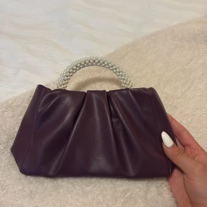 Liten lila handväska med pärlhandtag - Säljer en snygg lila handväska i skinnimitation med ett unikt handtag täckt av pärlor. Väskan har en cool veckad design och passar perfekt när du vill sticka ut lite extra. Perfekt accessoar för att lyfta din outfit!