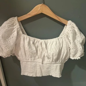 Vit croppad blus med puffärm - Supersöt vit croppad blus med broderade hål och puffiga korta ärmar. Toppen har smockad midja och elastisk halsringning som kan bäras offshoulder. Perfekt för sommarens alla tillfällen och ger en romantisk vibe.