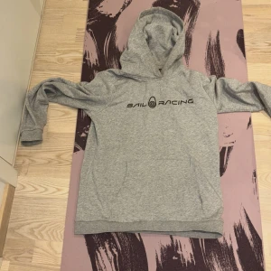 Grå hoodie från Sail Racing - Snygg grå hoodie från Sail Racing med svart logga på bröstet. Klassisk modell med huva och känguruficka framtill. Perfekt för chill dagar eller när du vill ha en sportig look. Mjuk och skön bomullsblandning, långärmad och med ribbade muddar.