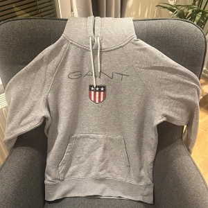 Grå hoodie från GANT (PRISET KAN DISKUTERAS VID SNABB AFFÄR) - Snygg grå hoodie från GANT med broderad logo och amerikanskinspirerat emblem på bröstet. Tröjan har huva med snörning och en stor magficka framtill. Perfekt för en chill och sportig look. Den är knappt använd och är i nyskick.