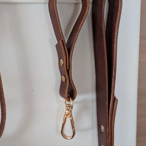 Brun nyckelring i skinn med gulddetaljer - Snyggt brun nyckelring i äkta skinn med stilrena guldfärgade metalldetaljer och nitar. Bältet har en enkel och klassisk design med karbinhake i metall, perfekt för att ge din outfit en cool touch. Handgjort av mig