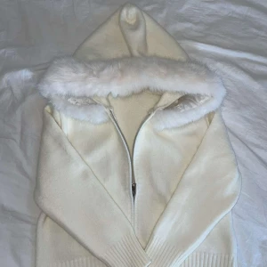 Vit stickad hoodie med fuskpäls - Mysig vit stickad hoodie med dragkedja och fluffig fuskpäls runt huvan. Jackan har långa ärmar, breda ribbade muddar och en clean look som passar perfekt till lager-på-lager-stil. Perfekt för kalla dagar när du vill vara både varm och snygg.