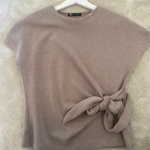 Beige stickad topp med knyt från Zara - Superfin beige stickad topp från Zara med korta ärmar och rund hals. Toppen har en snygg knytdetalj i sidan som ger en cool och trendig look. Perfekt att styla med jeans eller kjol för en chill vibe.