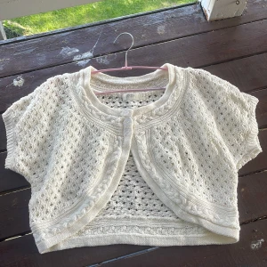 Vit stickad kort kofta från Jensen - Supersöt kort kofta från Jensen i vit stickad bomull. Koftan har korta ärmar, fina flätade detaljer längs kanterna och två knappar framtill. Perfekt att slänga över en klänning eller topp för en extra touch.